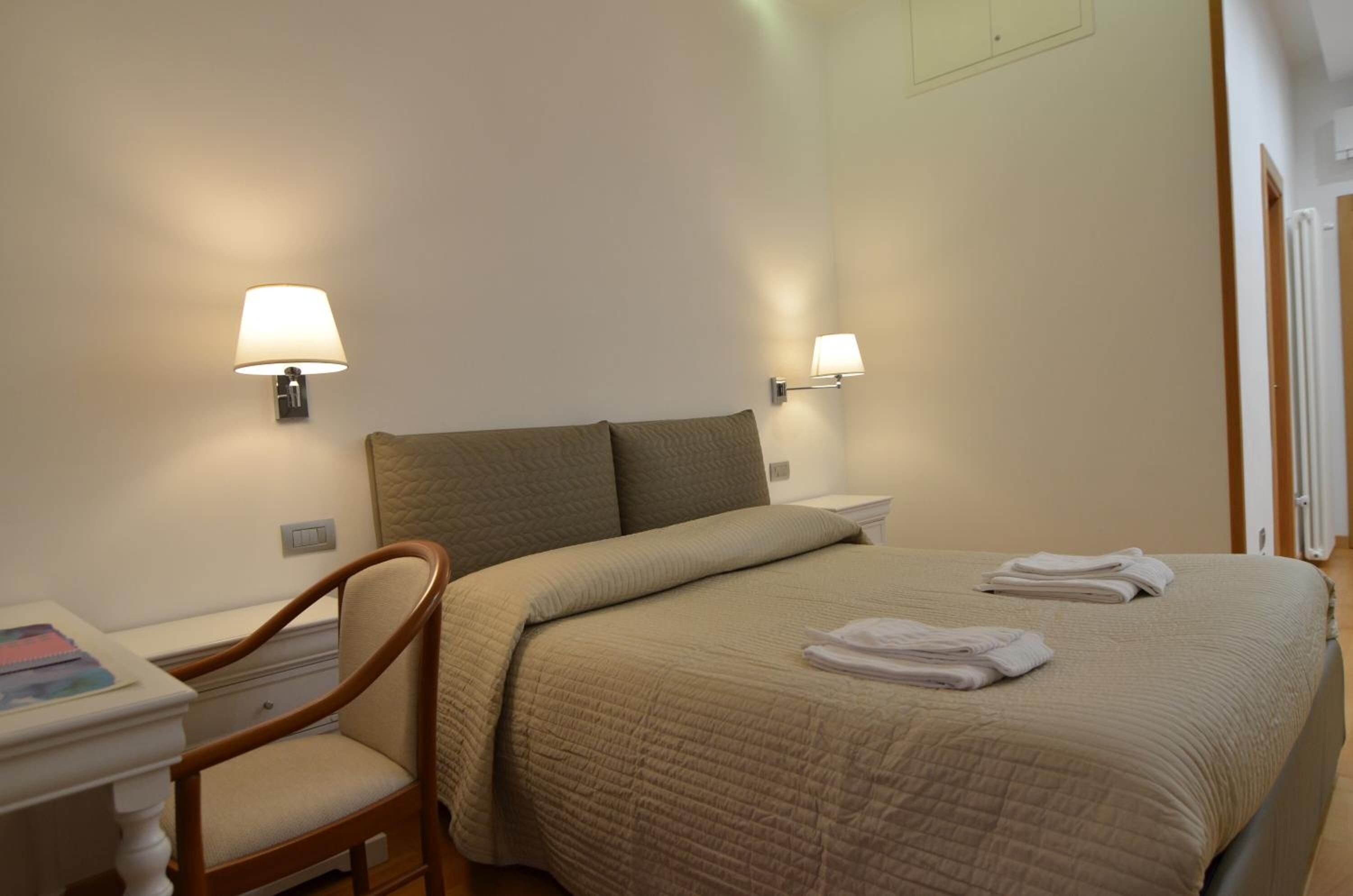 Een hotelkamer met een bed, twee bedlampen, een stoel en een klein tafeltje met een boek.