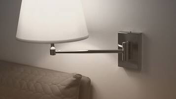 A wall-mounted lamp with a white shade and a metallic base, positioned next to a textured wall and a bed with a quilted cover.