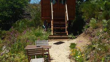 Una cabaña de madera con una escalera que lleva a una puerta, rodeada por un camino de grava y diversas plantas.