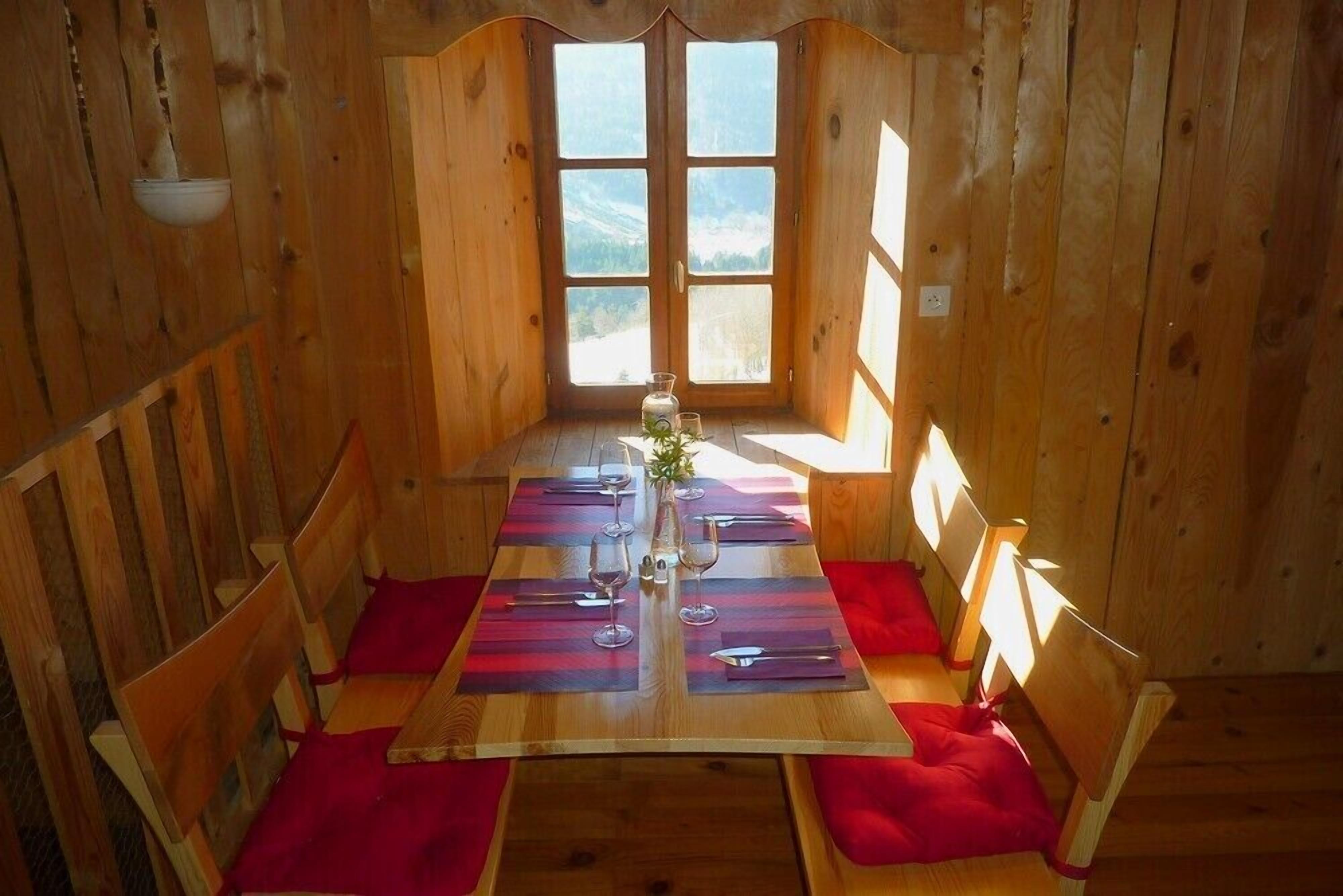 A wooden dining room with a table set for four, red cushions, and a view of the mountains through large windows.