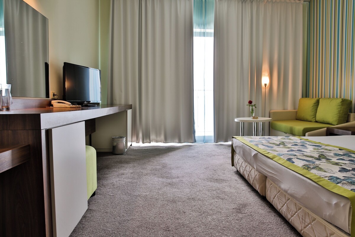 A hotel room with a bed, a desk with a television, a small table with a vase, and a window with curtains.