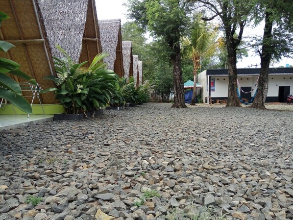 A courtyard with a gravel ground, potted plants, trees, and a building with a thatched roof. - Penginapan Bunar Tunggal (Tanjungjaya)