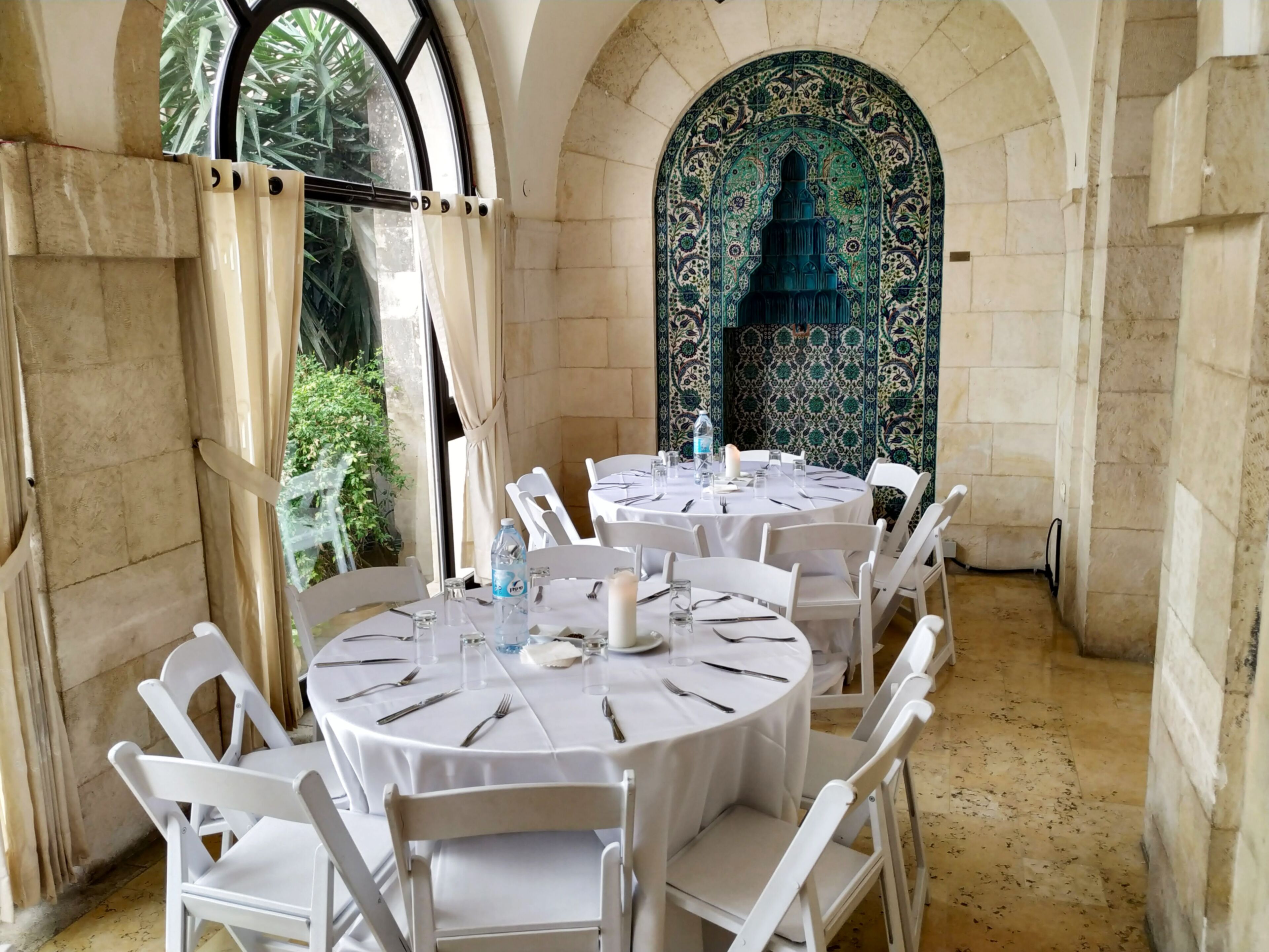 A dining area with round tables and white chairs, set against a backdrop of ornate tile work and a view of greenery outside.