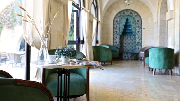 Un comedor con una mesa dispuesta para dos personas, un florero con cañas secas, una planta pequeña en maceta y un menú. La habitación tiene sillas tapizadas de color verde, un papel tapiz estampado y un suelo de baldosas.