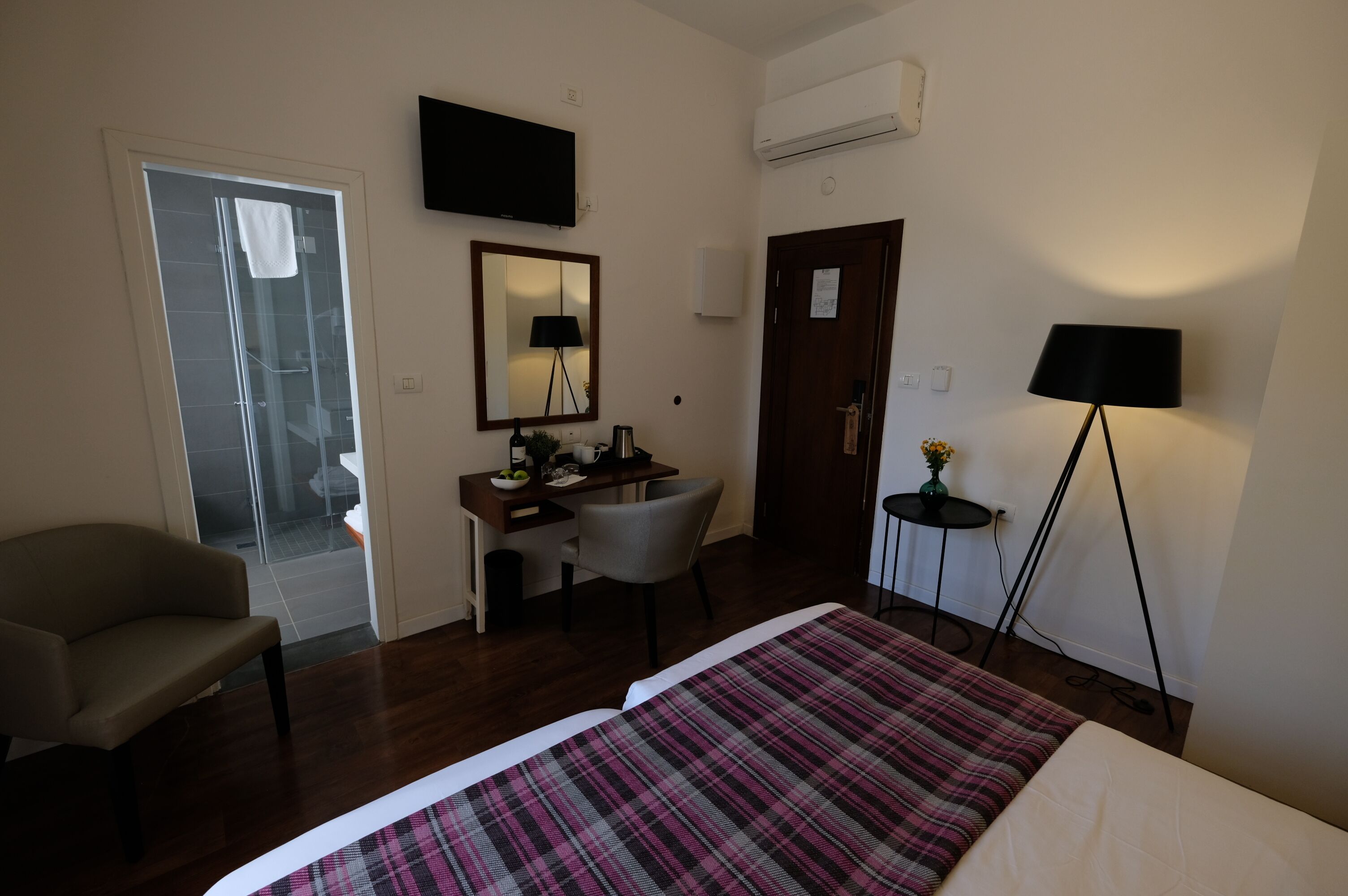 A hotel room with a bed, a desk with a chair, a TV, an air conditioner, and a lamp.