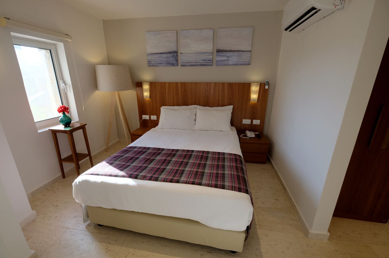 Un dormitorio con cama, mesitas de noche, lámpara, ventana y una obra de arte en la pared.
