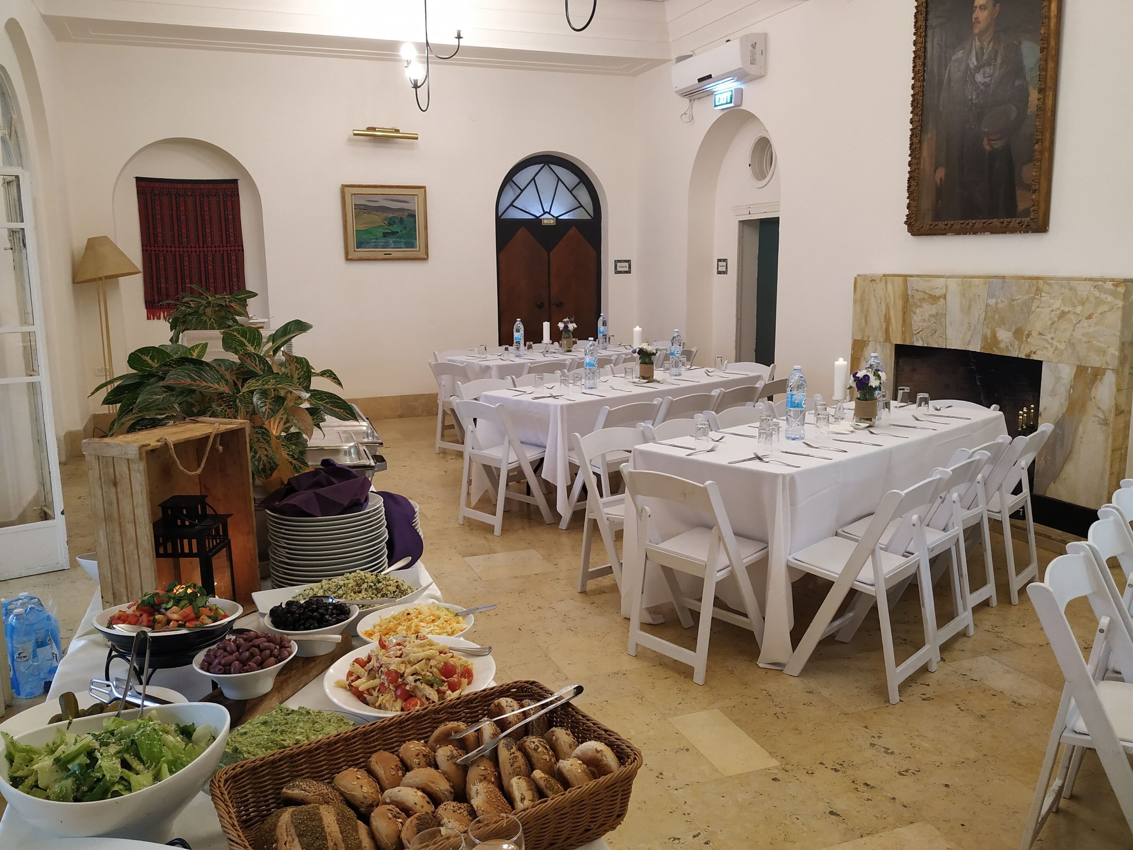 A dining hall with round tables set for a meal, white chairs, and a buffet with various dishes.