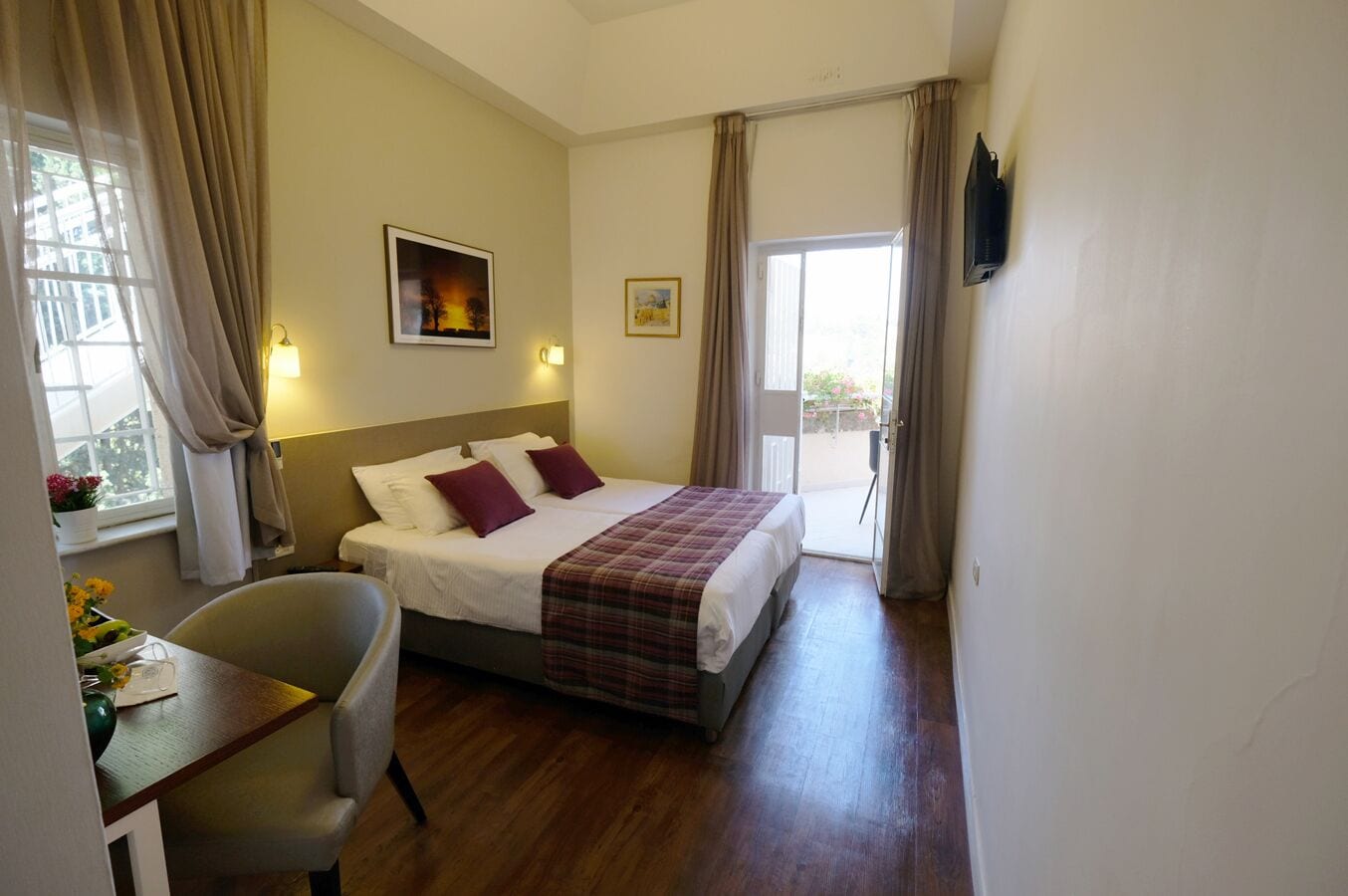 A hotel room with a bed, a desk, a chair, a window with curtains, and a door leading to an outdoor area.