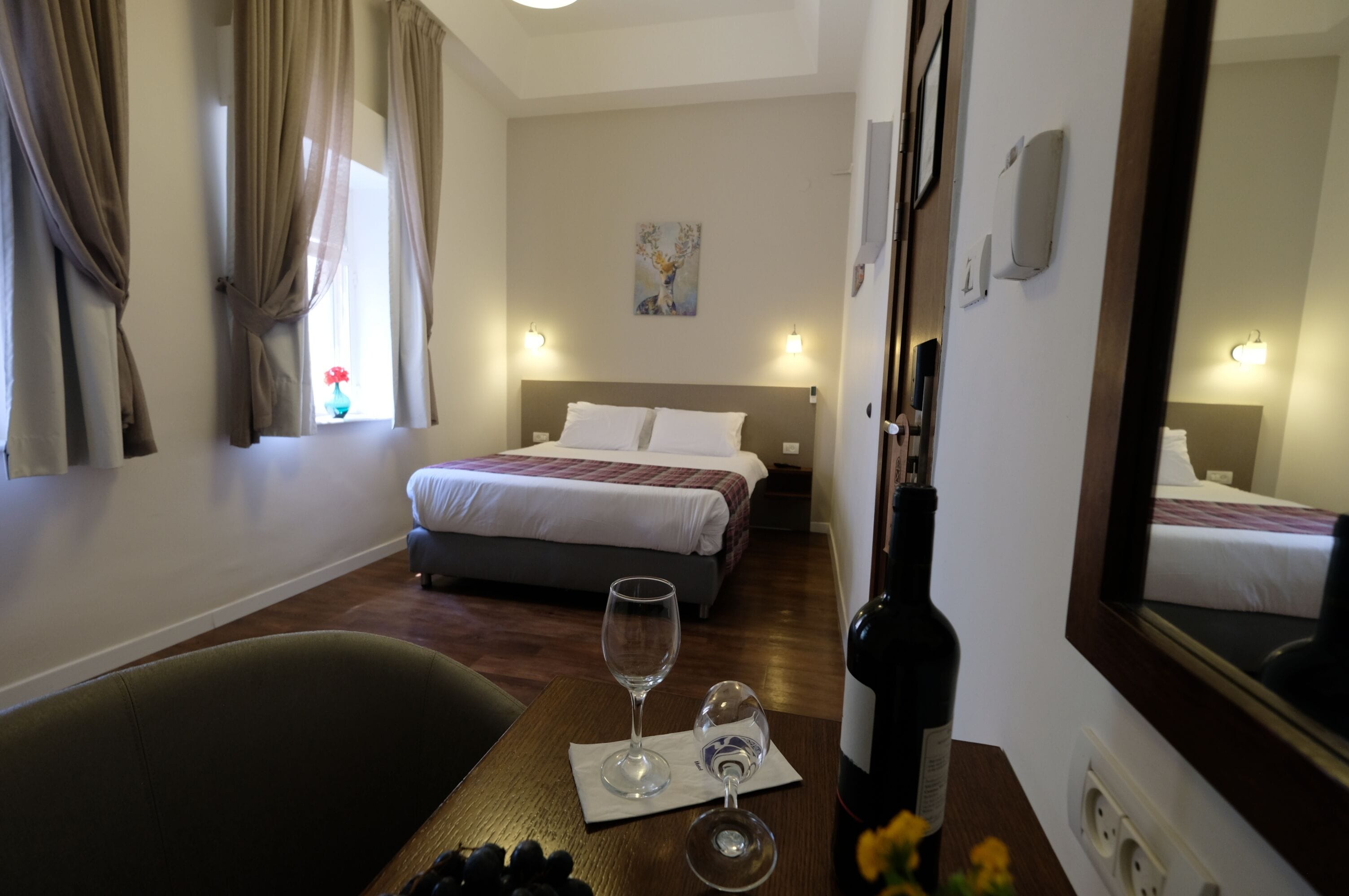 A hotel room with a bed, a table with a bottle and glasses, and a large mirror.