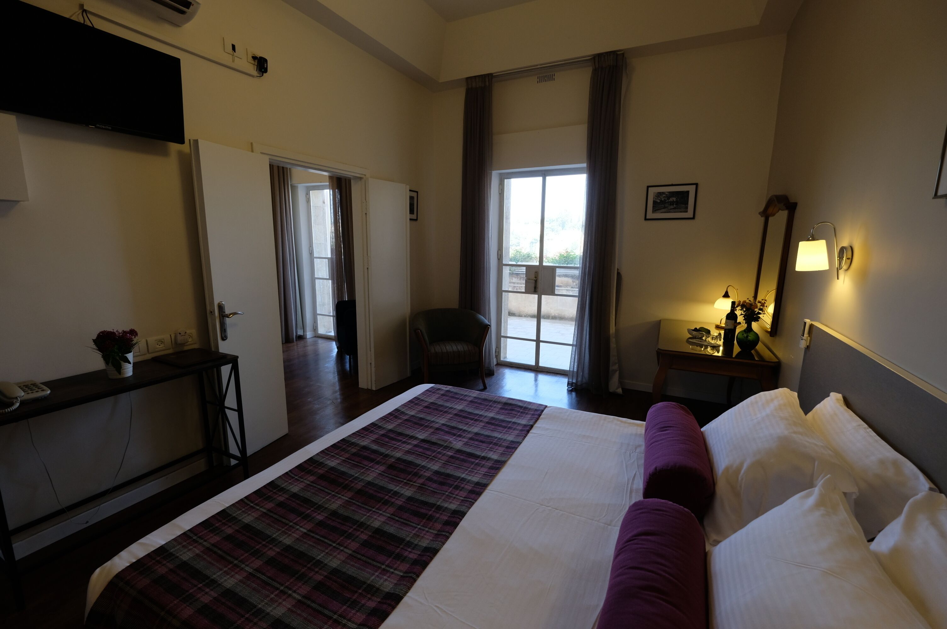 A hotel room with a large bed, a desk with a telephone, a chair, a window with curtains, and a small table with a lamp.