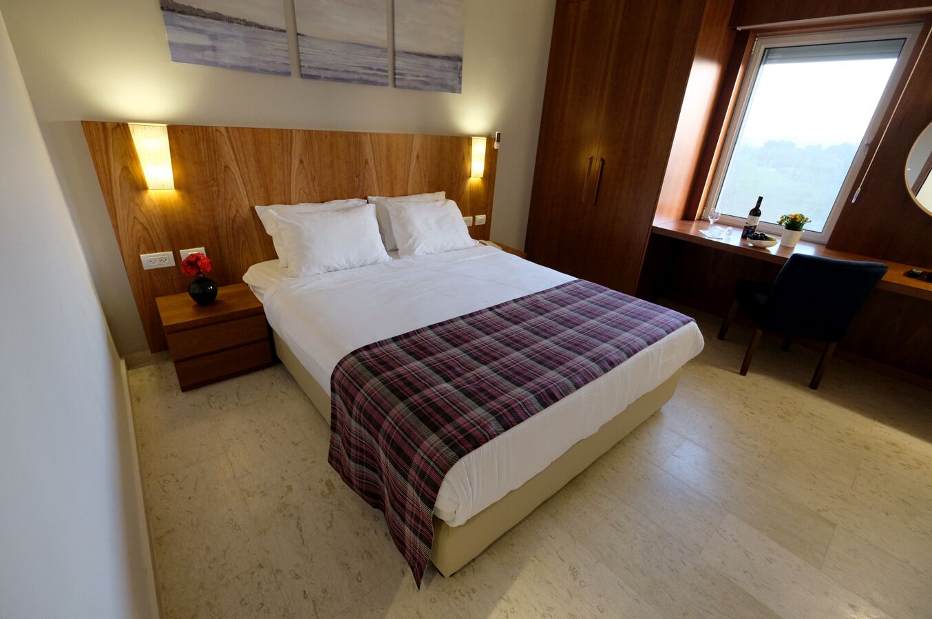 A bedroom with a large bed, wooden headboard, bedside lamps, a desk with a chair, and a window with a view.