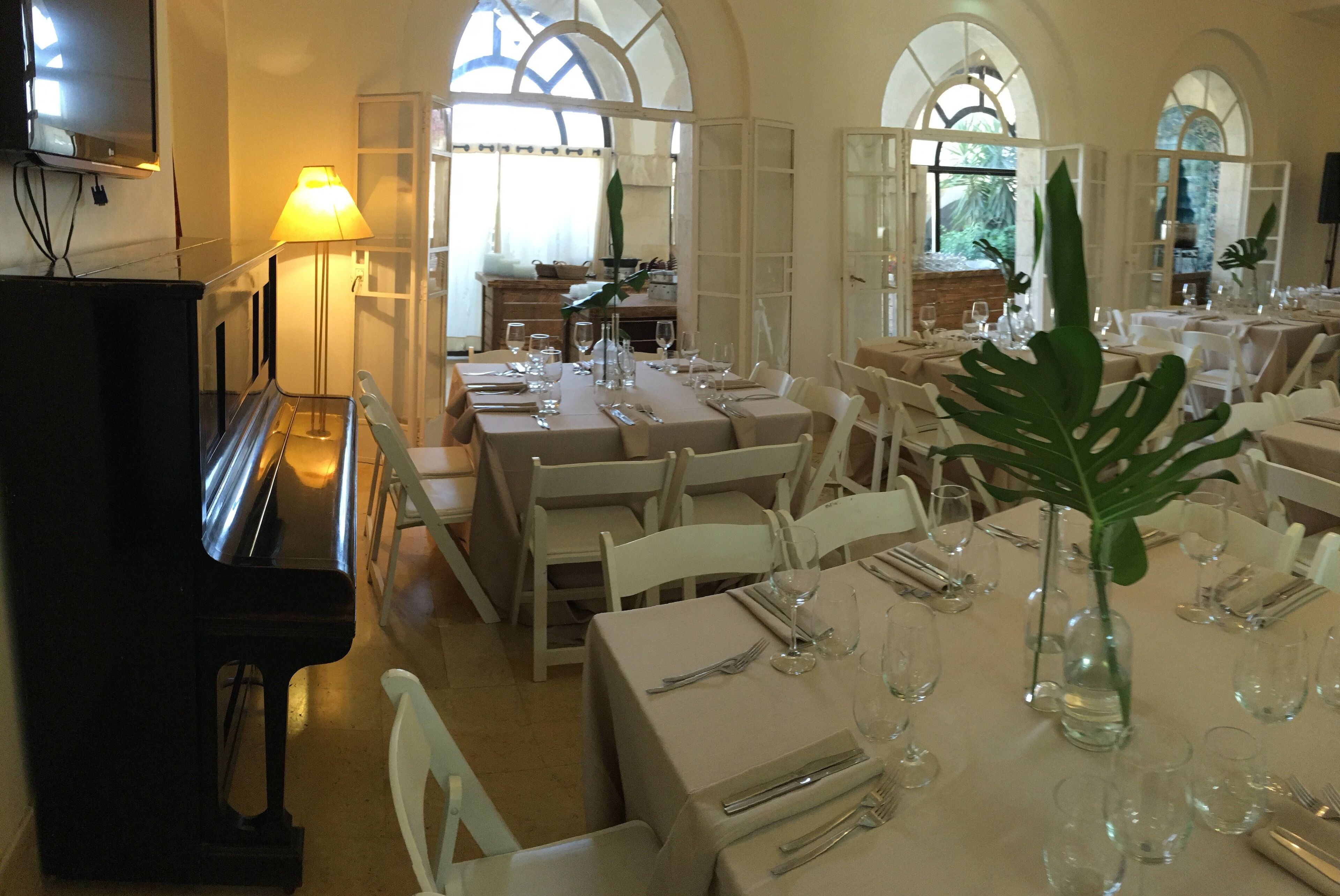 A dining area with tables set for a meal, a piano, and large windows with arched tops.