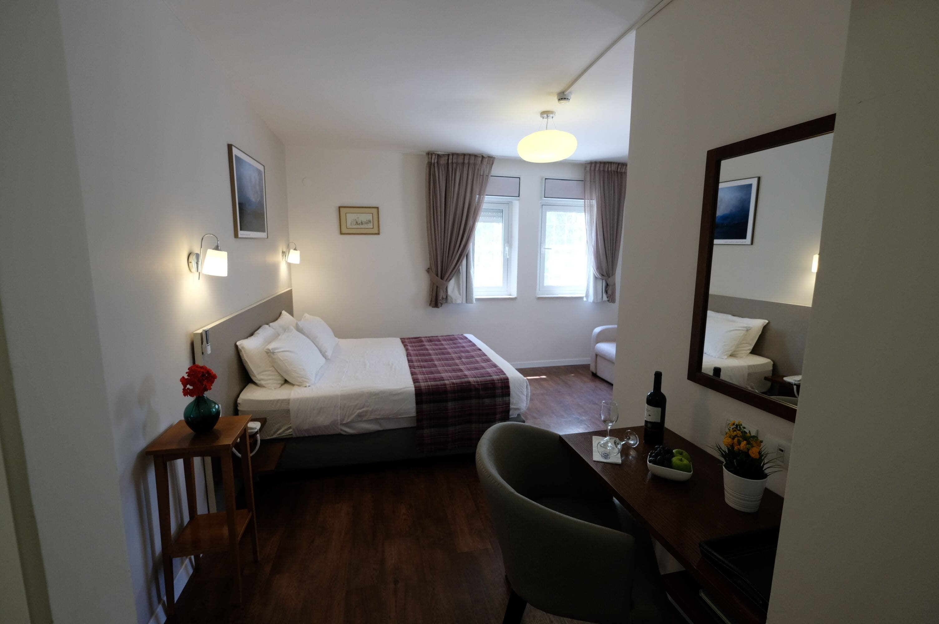 A hotel room with a bed, bedside table, chair, and a desk with a mirror. There are windows with curtains, a wall-mounted light, and a framed picture.