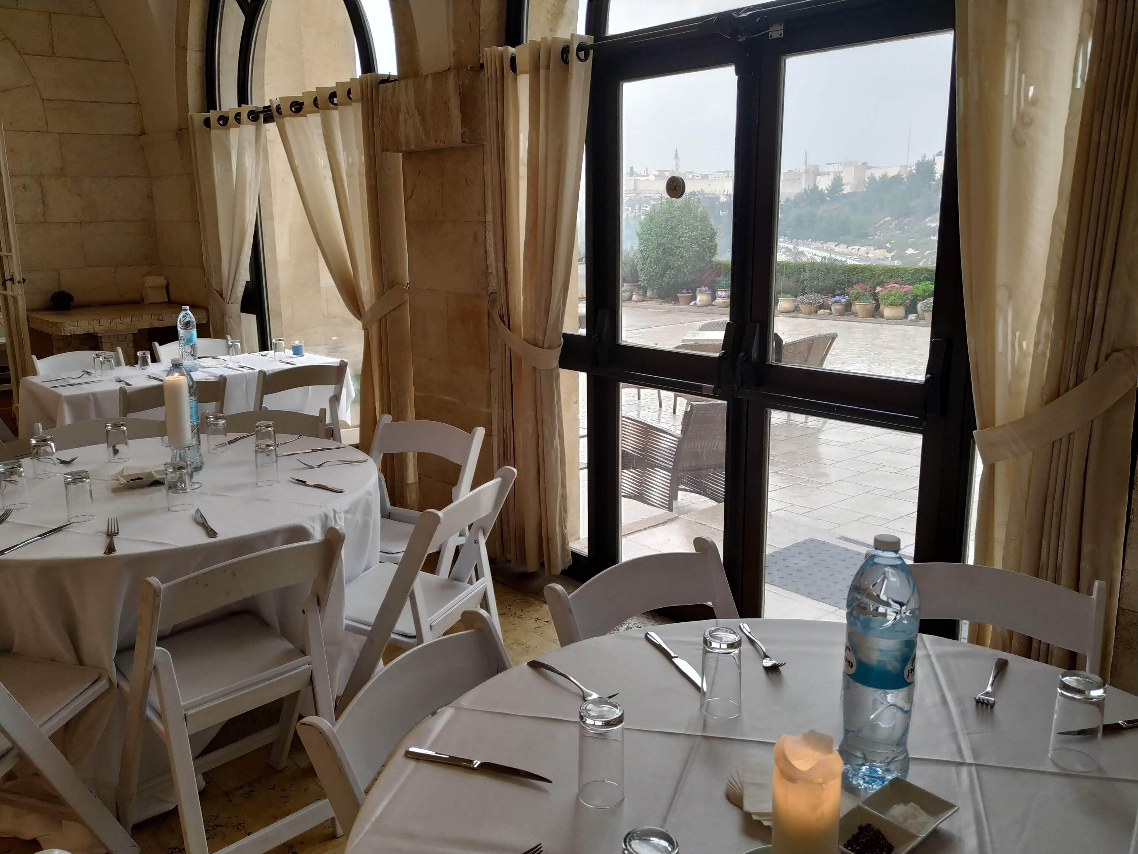 Un comedor con mesas redondas dispuestas para cuatro, cada una con mantel blanco, velas y cubiertos. Un amplio ventanal ofrece vistas de la ciudad.