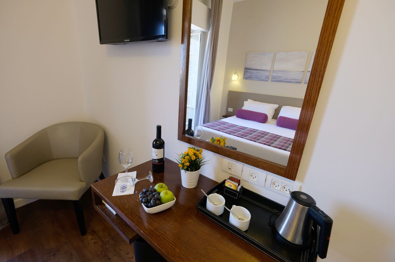 A hotel room with a bed, a TV mounted on the wall, a desk with a bottle of wine, a bowl of fruit, and a coffee maker.