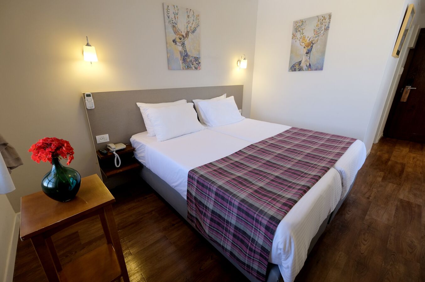 Une chambre d’hôtel avec un grand lit, deux lampes fixées au mur, une table de chevet ornée d’un vase de fleurs et deux tableaux accrochés au mur.