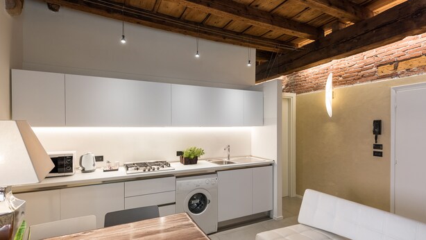 Eine moderne Küche mit weißen Schränken, einem eingebauten Geschirrspüler und einer Spüle. Im Wohnbereich befindet sich ein Holzesstzimmerstisch mit Stühlen sowie eine weiße Couch.