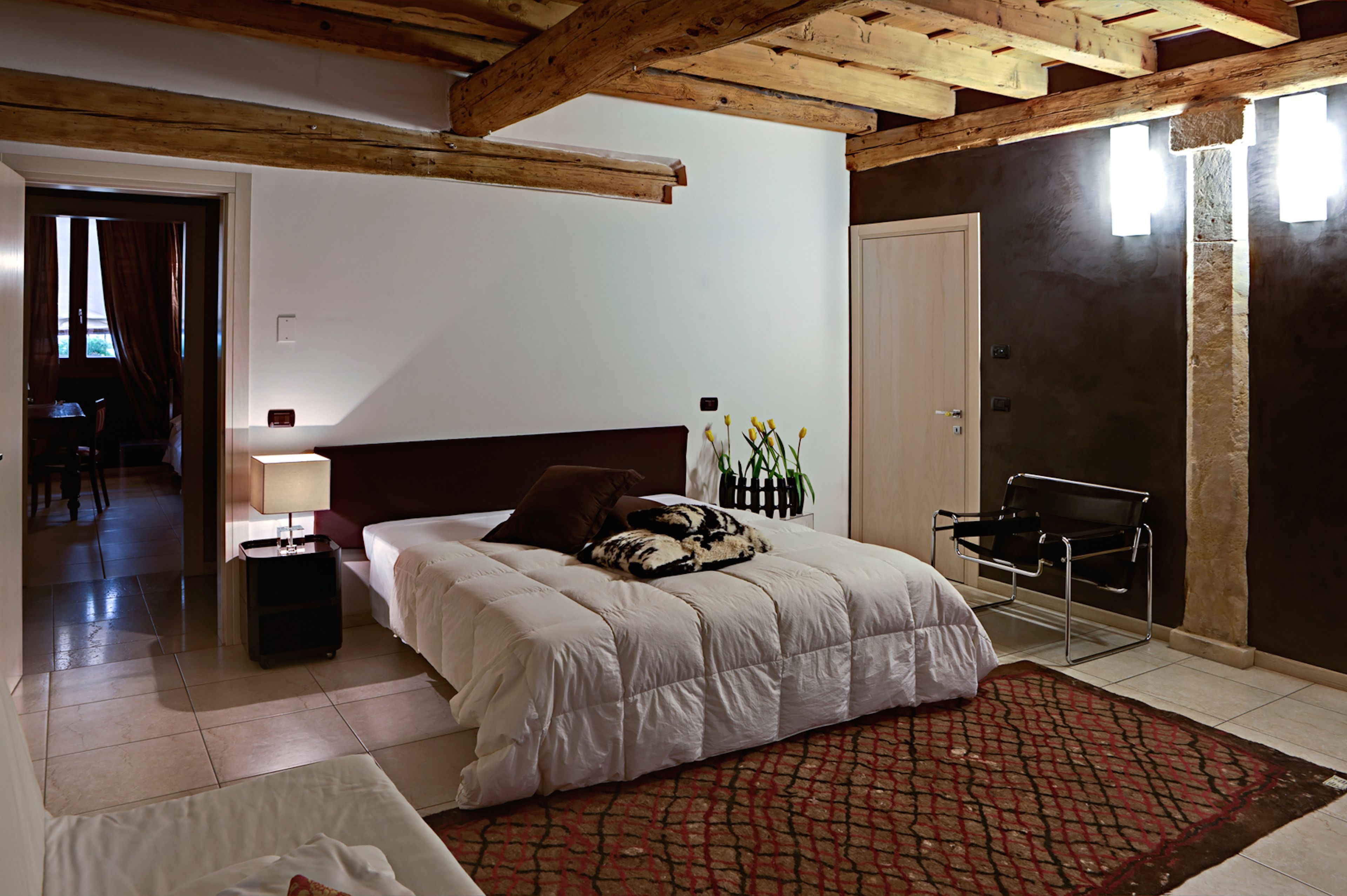Une chambre avec un grand lit, une table de chevet avec une lampe, un tapis rouge à motifs et une chaise noire.