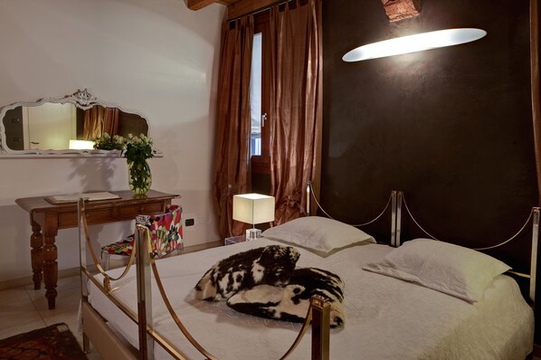 Un dormitorio con una cama con dosel, una mesita de noche con lámpara, un espejo y una ventana con cortinas.