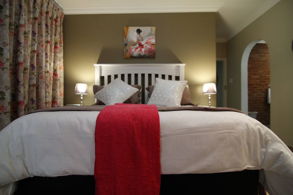 A neatly made bed with a red throw, two pillows, and a headboard. There is a painting above the bed and a floral curtain to the left.
