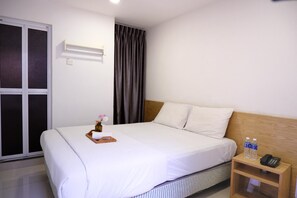 A hotel room with a bed, a bedside table with a phone and water bottles, a window with curtains, and a wall-mounted air conditioner. - Iris Hotel (Kajang)