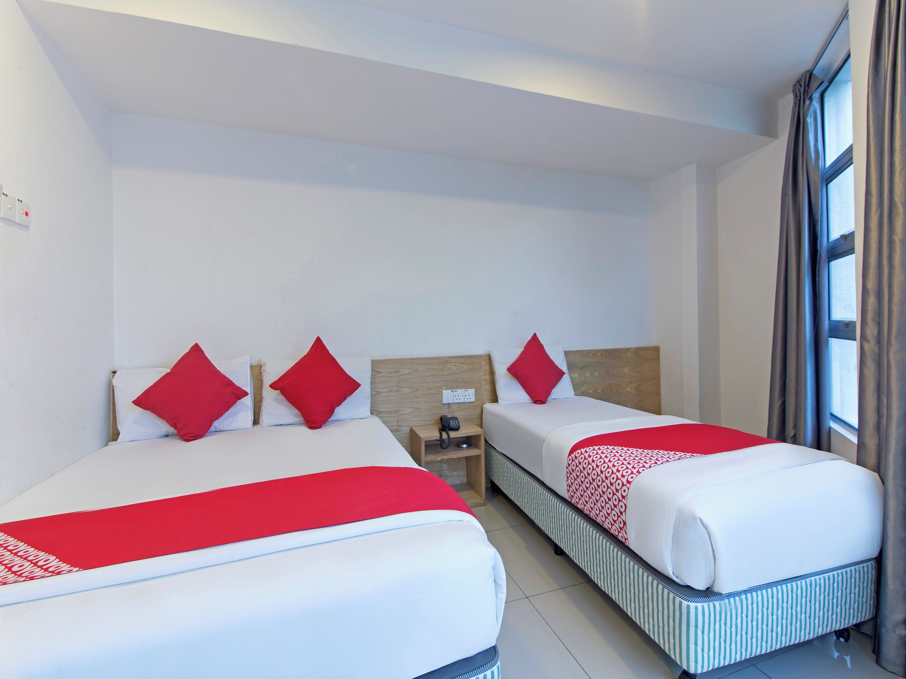 A hotel room with two beds, each with red pillows, a wooden headboard, a bedside table with a phone, and a window with curtains.