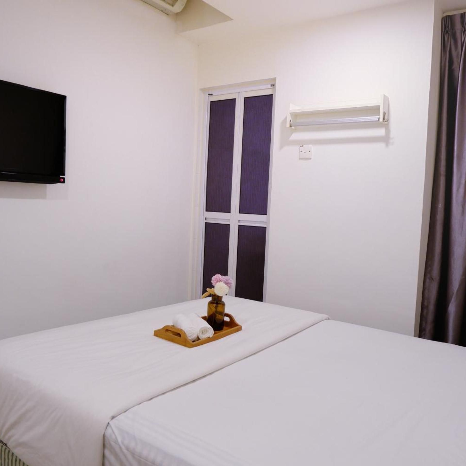 A hotel room with a bed, a TV mounted on the wall, a window with curtains, and a small table with towels and a vase.