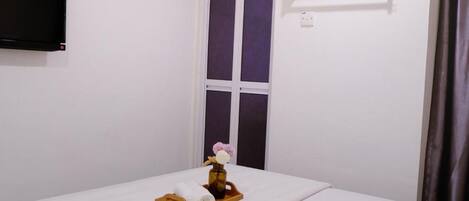 A hotel room with a bed, a TV mounted on the wall, a window with curtains, and a small table with towels and a vase.