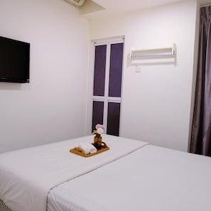 Ett hotellrum med en säng, en tv som är monterad på väggen, ett fönster med gardiner och ett litet bord med handdukar och en vas.