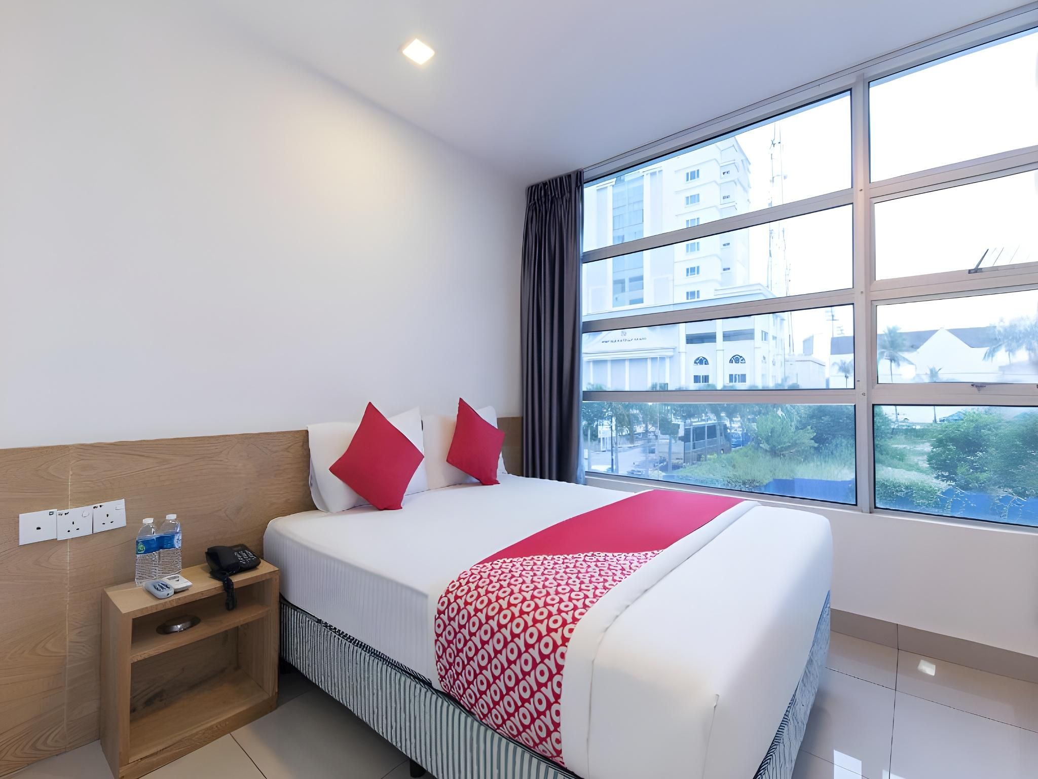 Une chambre d’hôtel avec un grand lit, une table de chevet avec un téléphone et des bouteilles d’eau, une prise électrique et une fenêtre donnant sur des immeubles et des arbres.