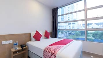 Une chambre d’hôtel avec un grand lit, une table de chevet avec un téléphone et des bouteilles d’eau, une prise électrique et une fenêtre donnant sur des immeubles et des arbres.