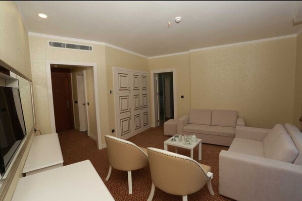 A living room with a beige sofa, two armchairs, a coffee table, and a television. There are doors leading to other rooms.
