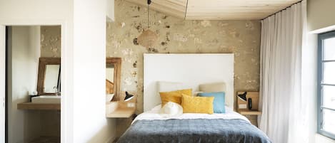 A bedroom with a large bed, a wooden ceiling, a stone wall, and a window with curtains.