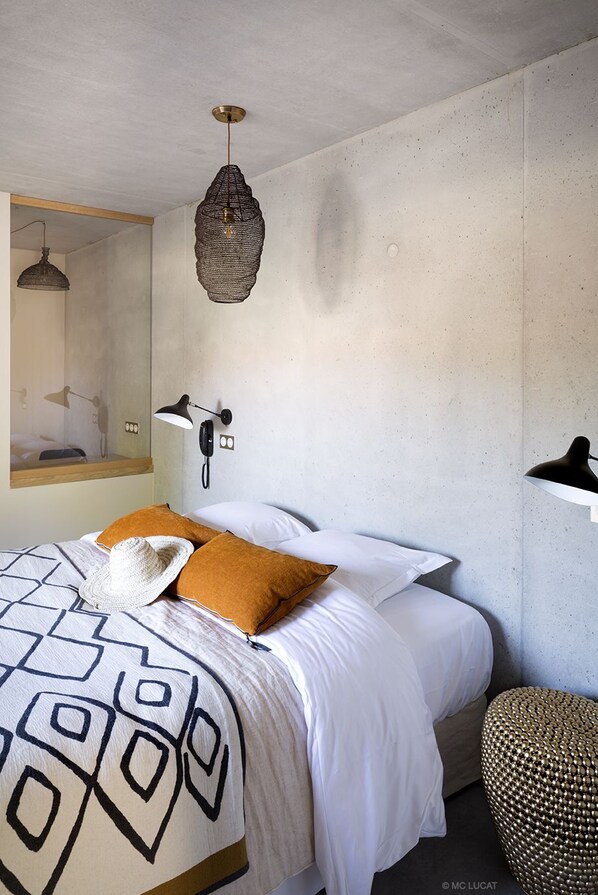 A bedroom with a bed, a hanging light fixture, a wall-mounted phone, and a small stool. - Hotel 19-21 (Leucate)