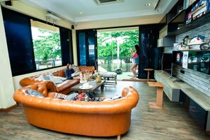 A modern living room with a large orange leather sofa, a wooden coffee table, and a small wooden bench. There is a balcony with a view of greenery outside. - Doolay Beachfront Hostel (Karon)