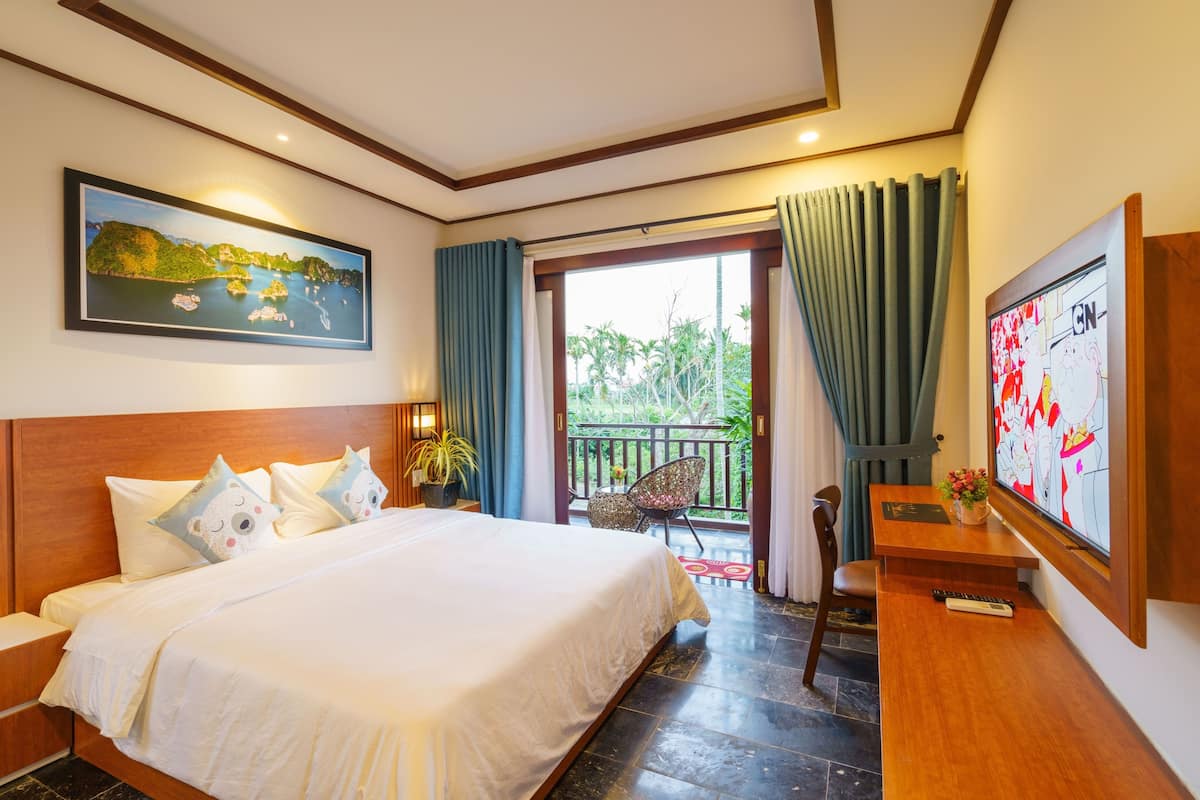 A hotel room with a large bed, a desk, a TV, a balcony with a view of greenery, and a painting on the wall.