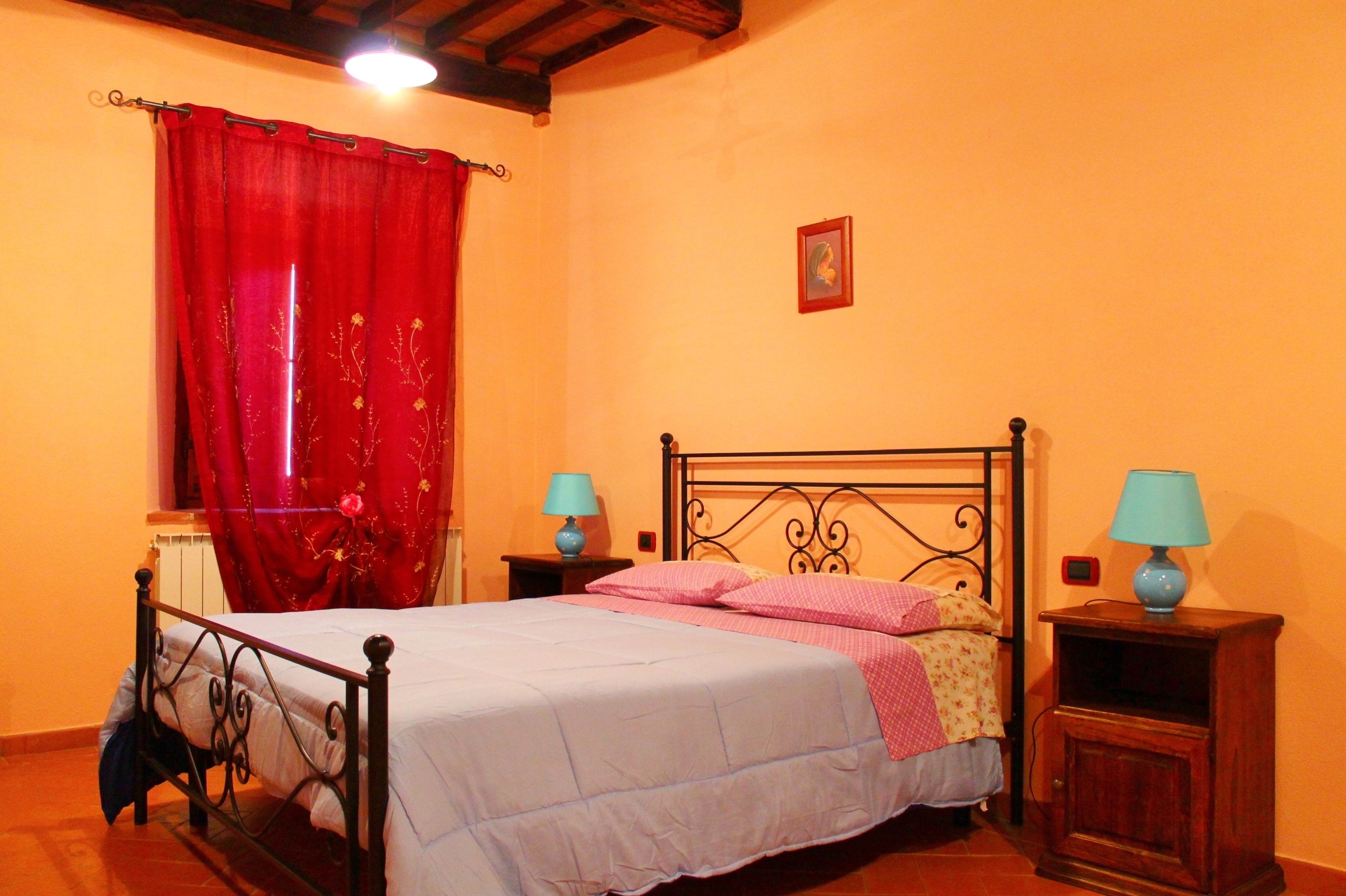 Un dormitorio con cama, cortinas rojas, dos mesitas de noche con lámparas, un radiador y un cuadro en la pared.
