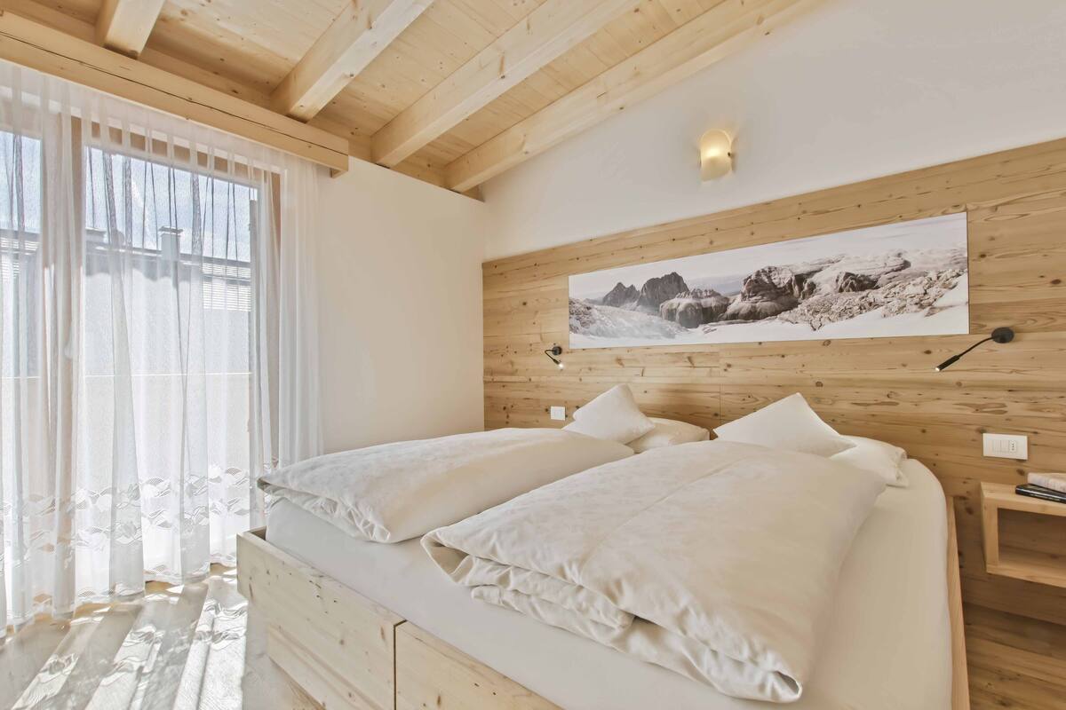 Un dormitorio con techo de madera, cama con ropa blanca, ventana grande con cortinas traslúcidas y un cuadro colgado en la pared.