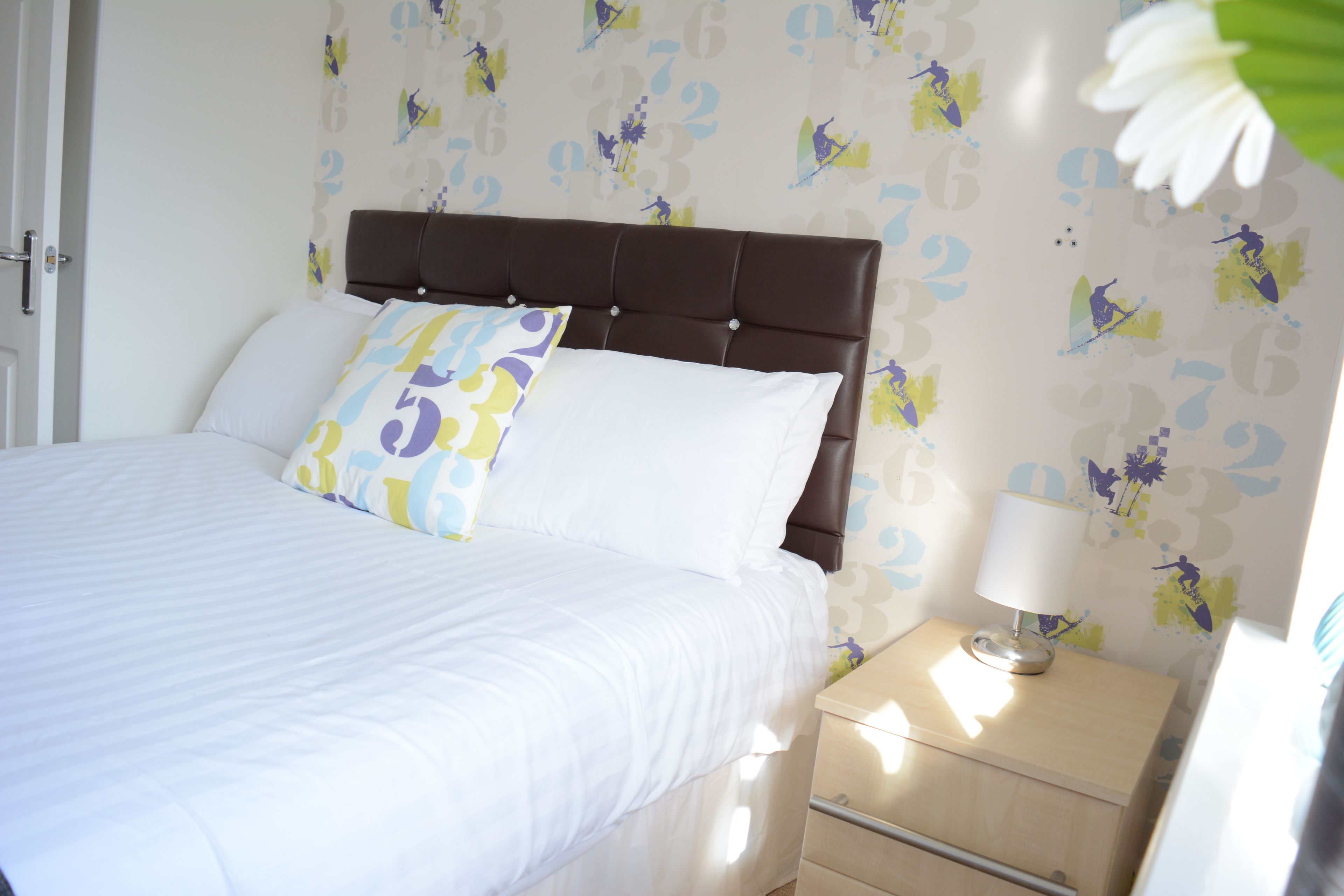 A neatly made bed with white bedding and a brown headboard, a bedside table with a lamp, and a wall with a floral pattern.