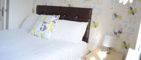 A neatly made bed with white bedding and a brown headboard, a bedside table with a lamp, and a wall with a floral pattern.