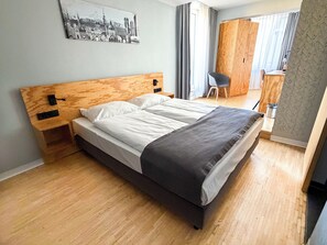 A modern hotel room with a wooden headboard, a large bed with white and gray bedding, a wooden nightstand, a desk with a chair, and a wardrobe. - mk | hotel münchen city (Munich)