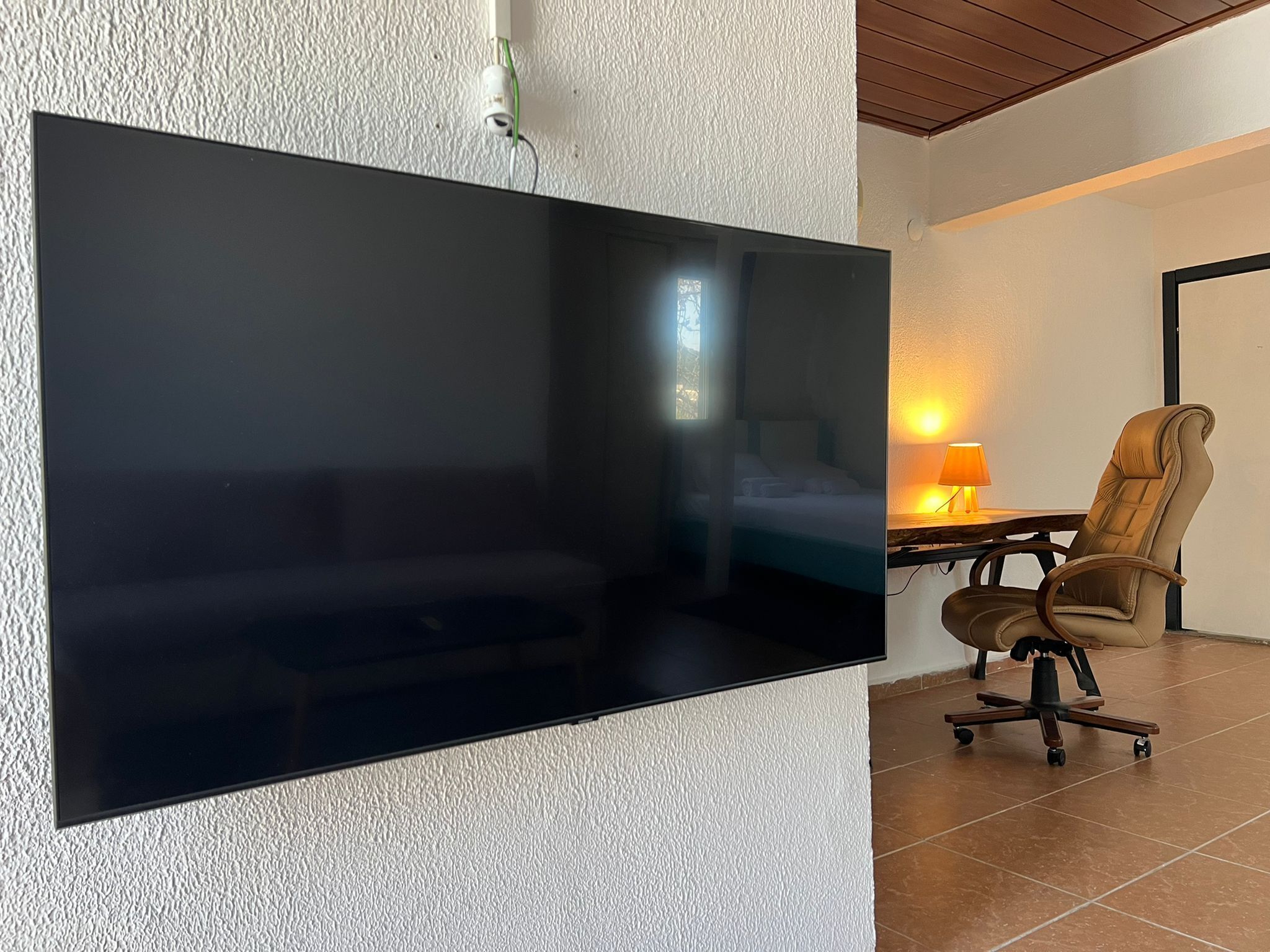 Un televisor de pantalla plana montado en una pared con textura, techo de madera, cama con cabecera blanca, lámpara y sillón de cuero marrón.