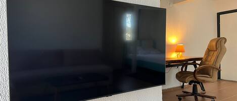 Un televisor de pantalla plana montado en una pared con textura, techo de madera, cama con cabecera blanca, lámpara y sillón de cuero marrón.