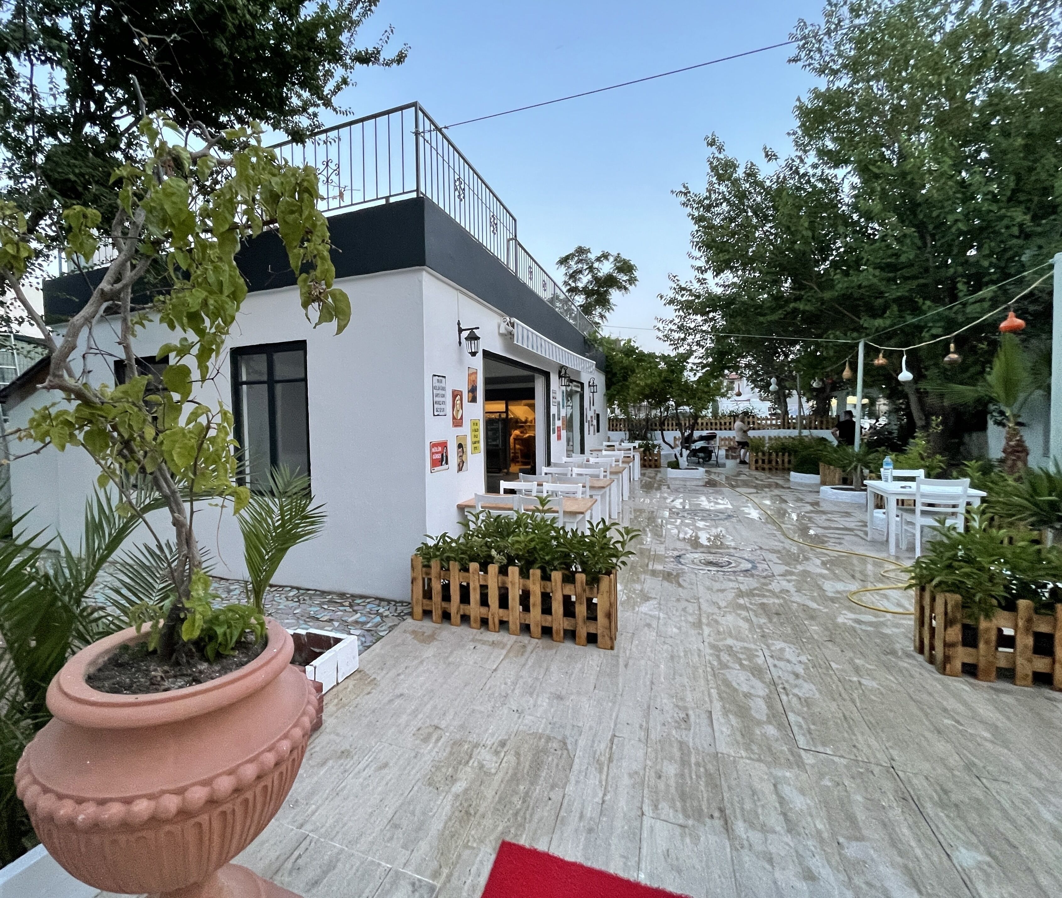 Un área de café al aire libre con mesas y sillas, un edificio blanco con techo negro y plantas en macetas.