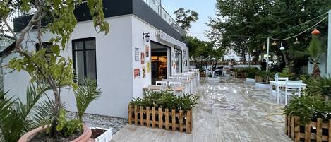 Un área de café al aire libre con mesas y sillas, un edificio blanco con techo negro y plantas en macetas.