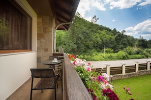 A balcony with wicker furniture, a stone wall, and a view of a road and forested hill. - Vitora Guest House (Teteven)