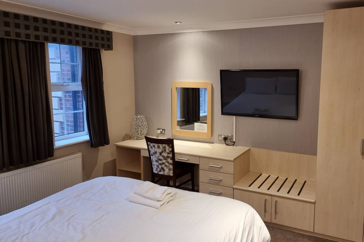 A hotel room with a bed, a desk with a mirror, a television, and a wardrobe.