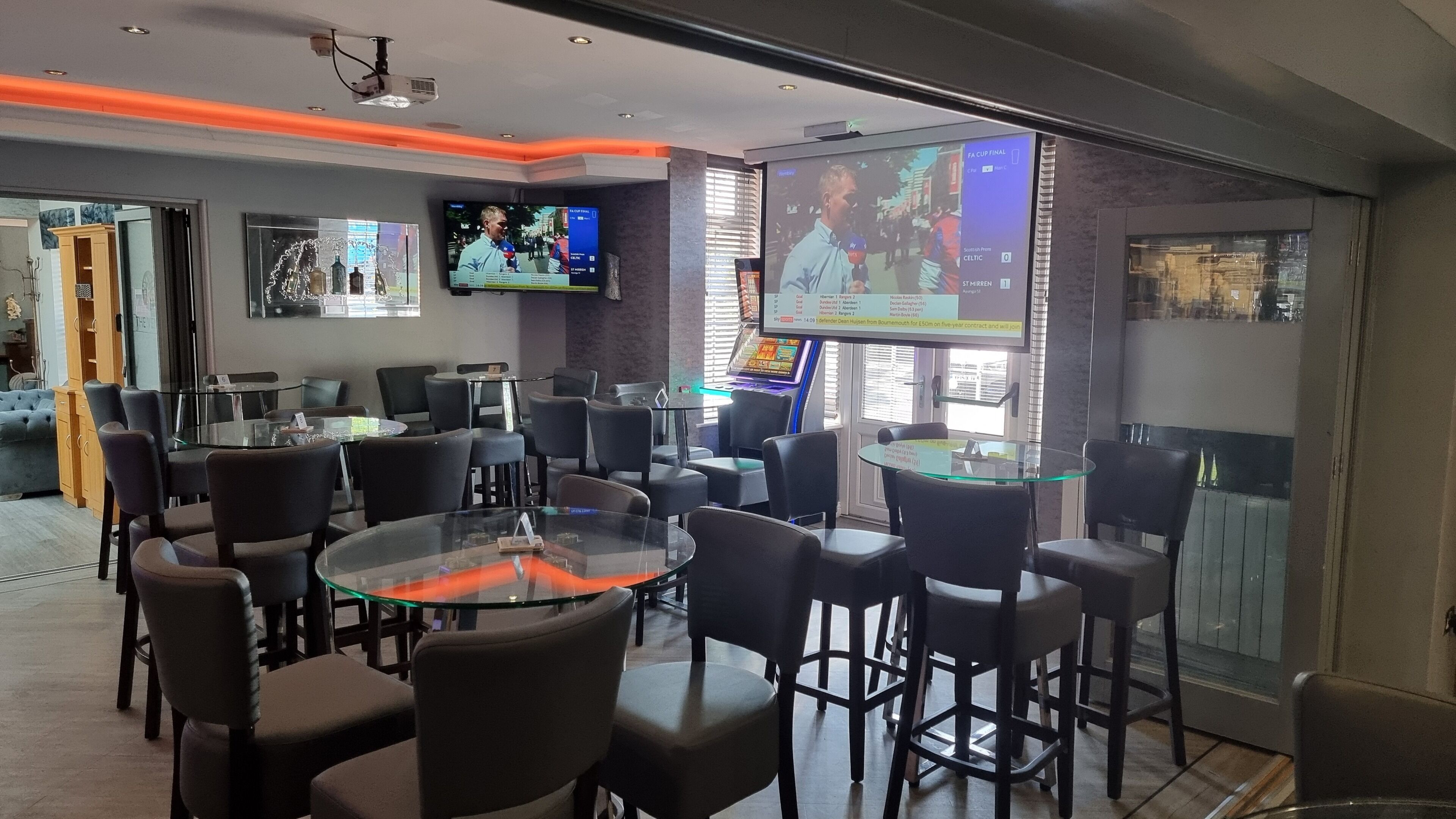 A modern bar area with multiple seating options, including round tables and bar stools. There are televisions mounted on the walls displaying sports events.