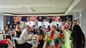Un evento concurrido en un espacio interior, con mesas, sillas, luces de techo y un proyector.