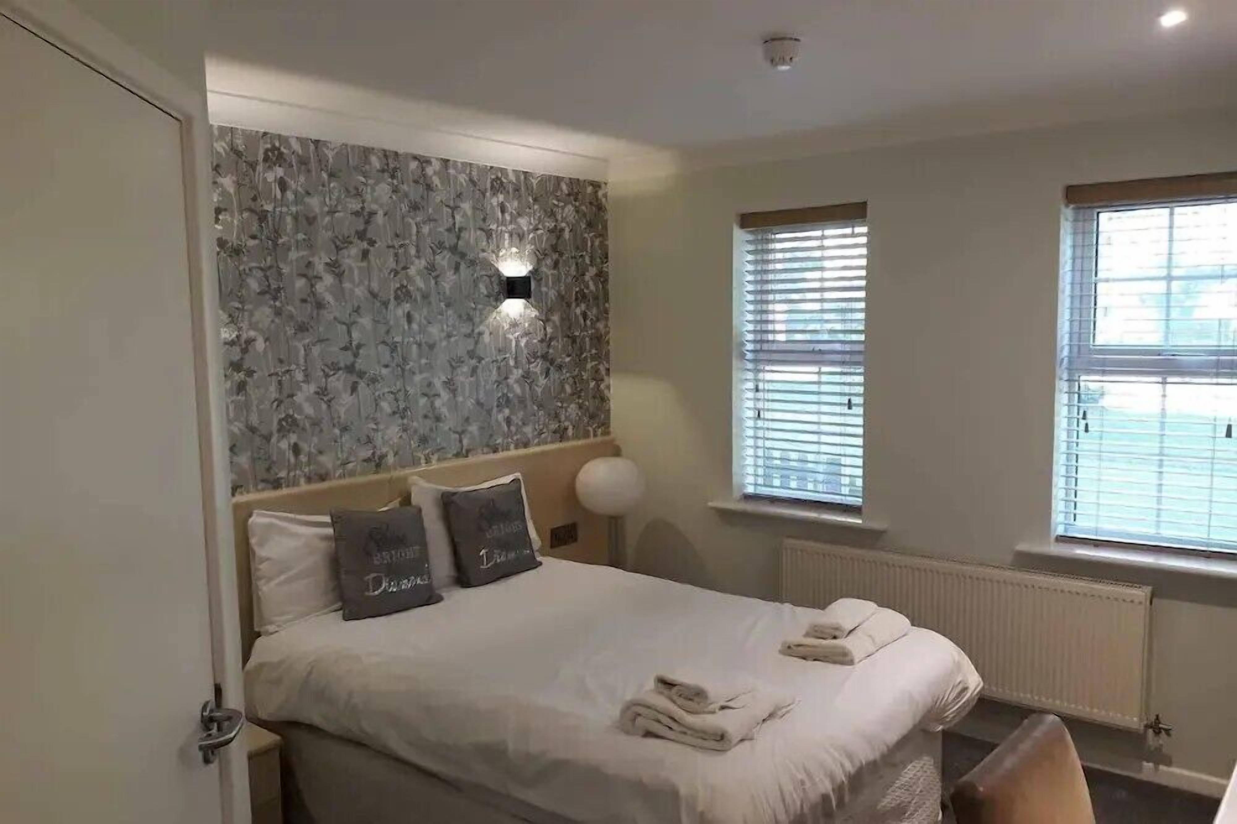 A bedroom with a bed, two pillows, a wall-mounted light, a window with blinds, and a radiator.
